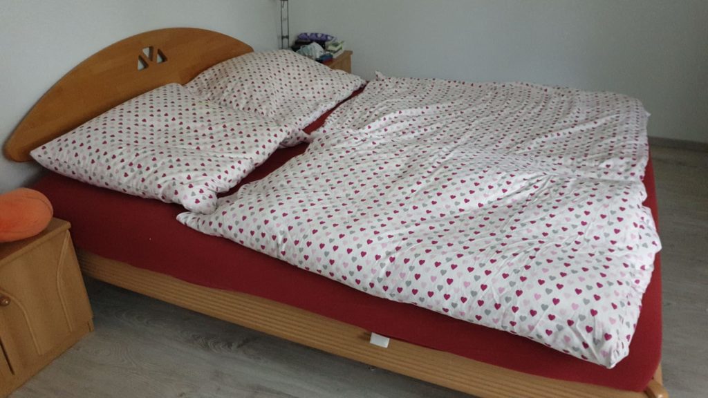 man sieht: großes Doppelbett aus angenehm braunem Buchenholz, darauf liegt eine rote Matratze und ein mit Herzen bedruckt, darunter sieht eine rote Matratze und eine mit verschiedenfarbigen Herzen bedruckte weise Bettwäsche